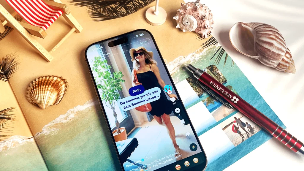 TikTok-Marketing für TUI Austria von der MMCAgentur: Ein Smartphone zeigt ein Urlaubsvideo inmitten von Muscheln, Sand und einem roten MMC-Kugelschreiber.