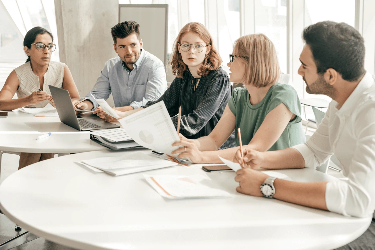 Eine Gruppe aus drei Frauen und zwei Männern sitzt mit Laptop und Ausdrucken an zwei runden Tischen und diskutiert.