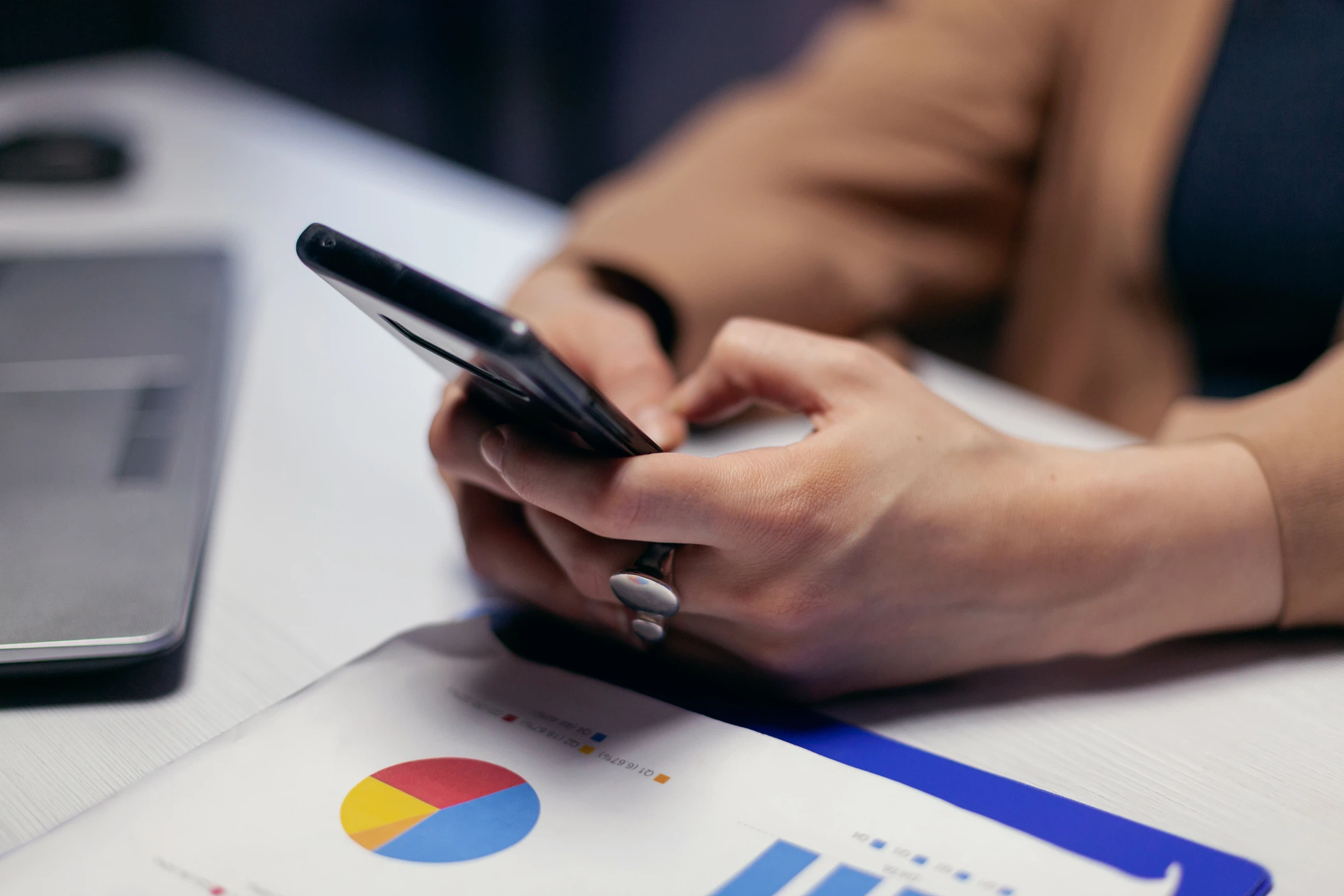 Digitale Analyse und Projektarbeit: Eine Person nutzt ein Smartphone neben einem Laptop und ausgedruckten Diagrammen und Statistiken.