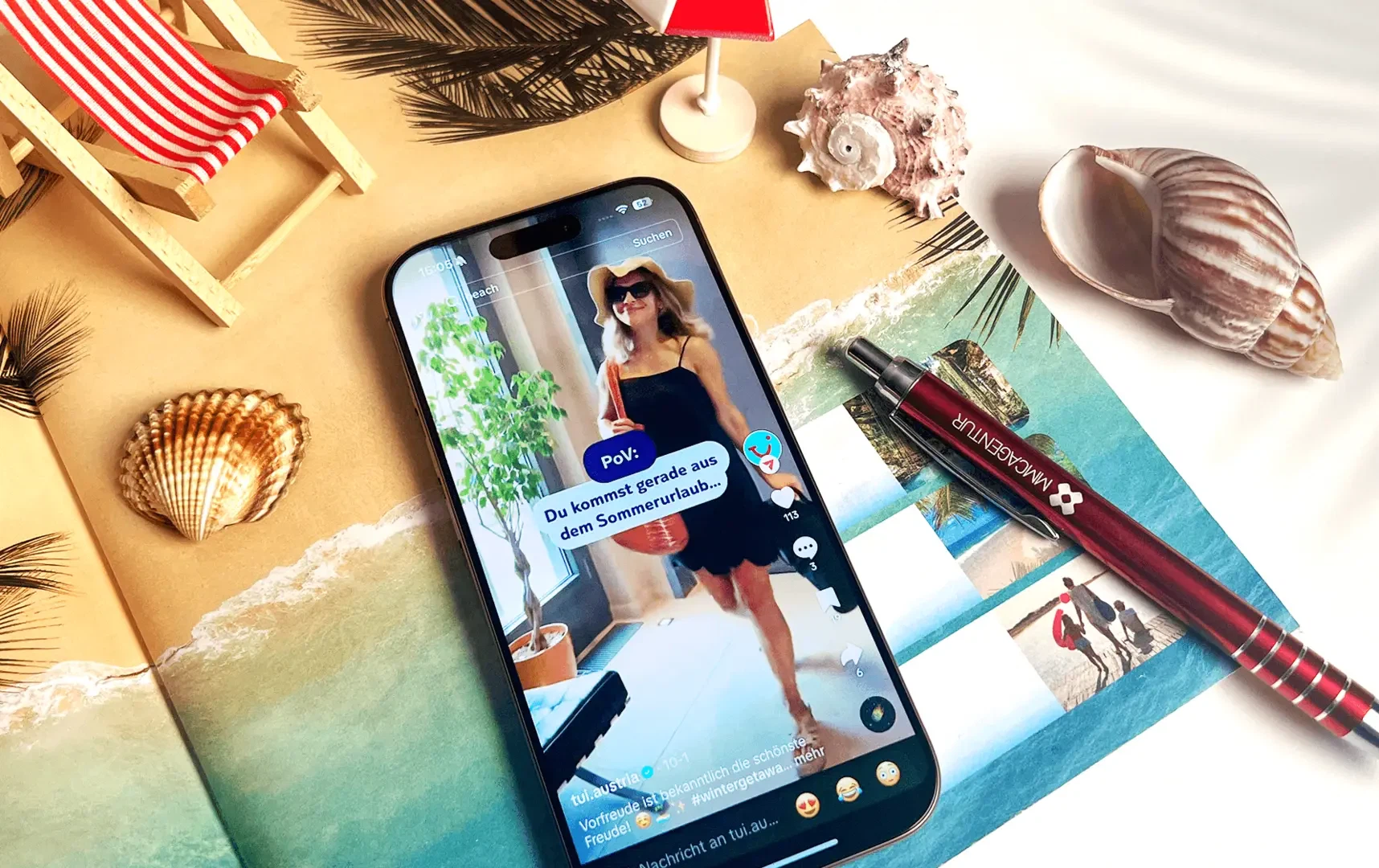TikTok-Marketing für TUI Austria von der MMCAgentur: Ein Smartphone zeigt ein Urlaubsvideo inmitten von Muscheln, Sand und einem roten MMC-Kugelschreiber.